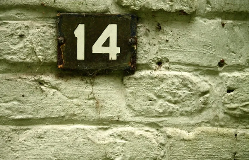A weathered metal plaque numbered 14 fixed to a rough painted brick wall