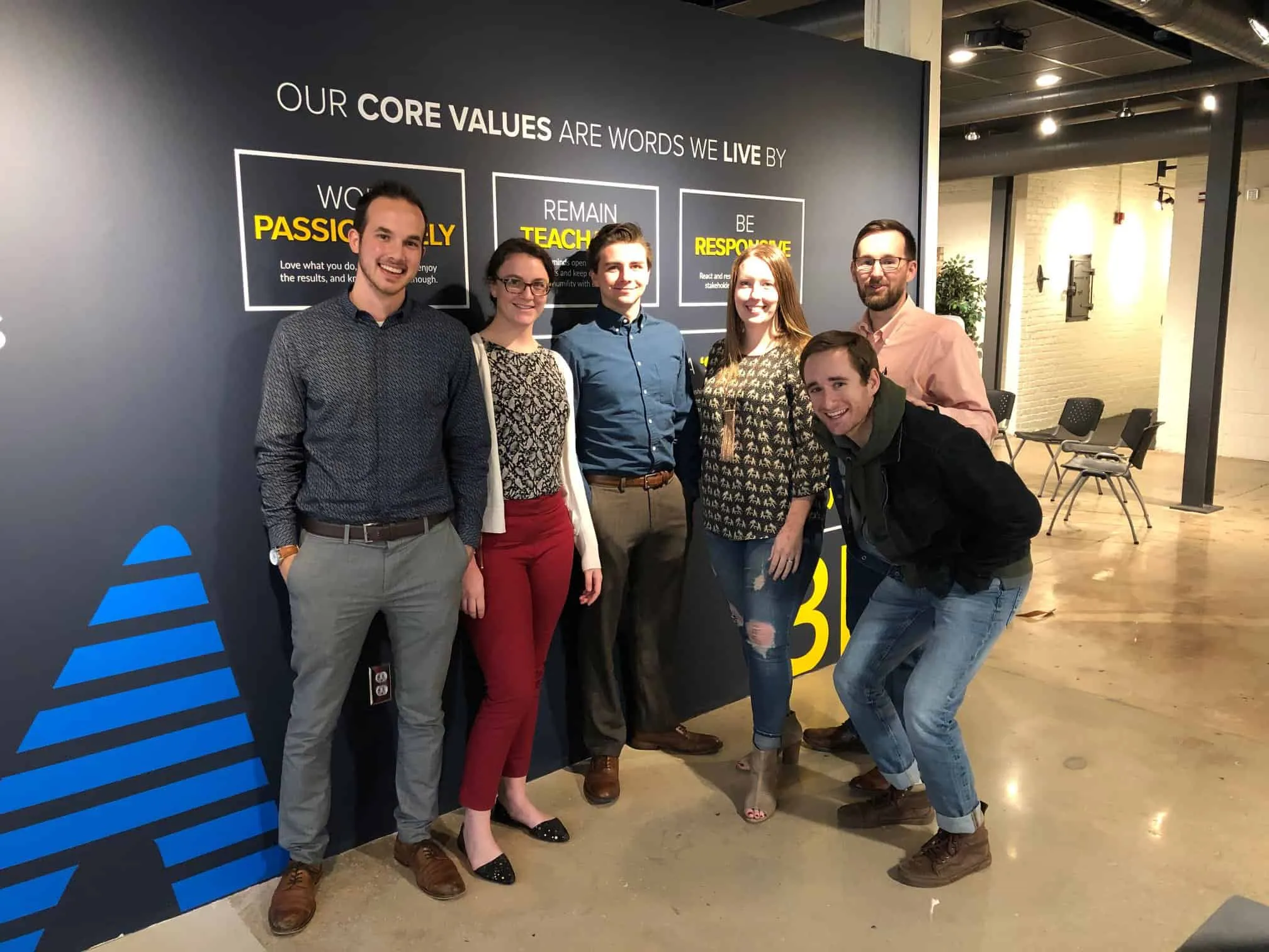 Six colleagues posing in front of an office wall that reads our core values are words we live by
