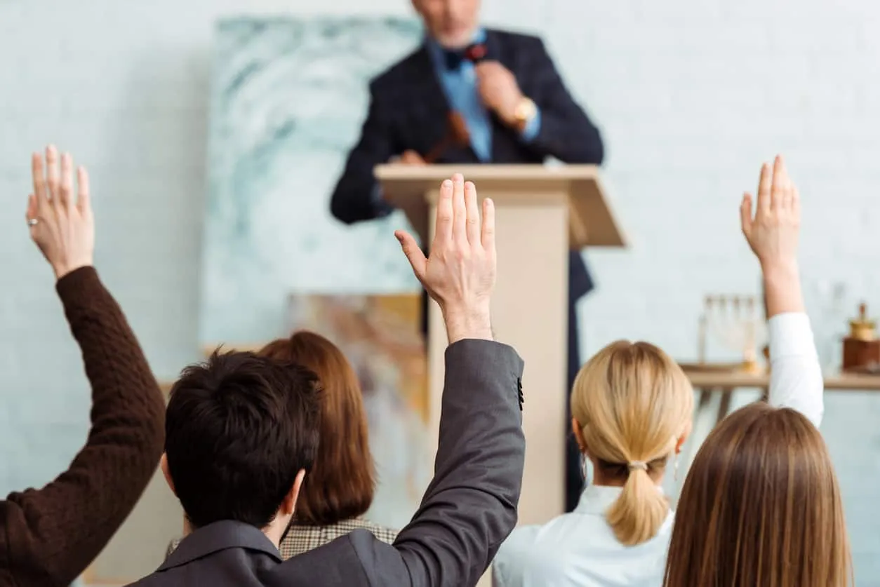 Raised hands in an audience facing a speaker at a lectern