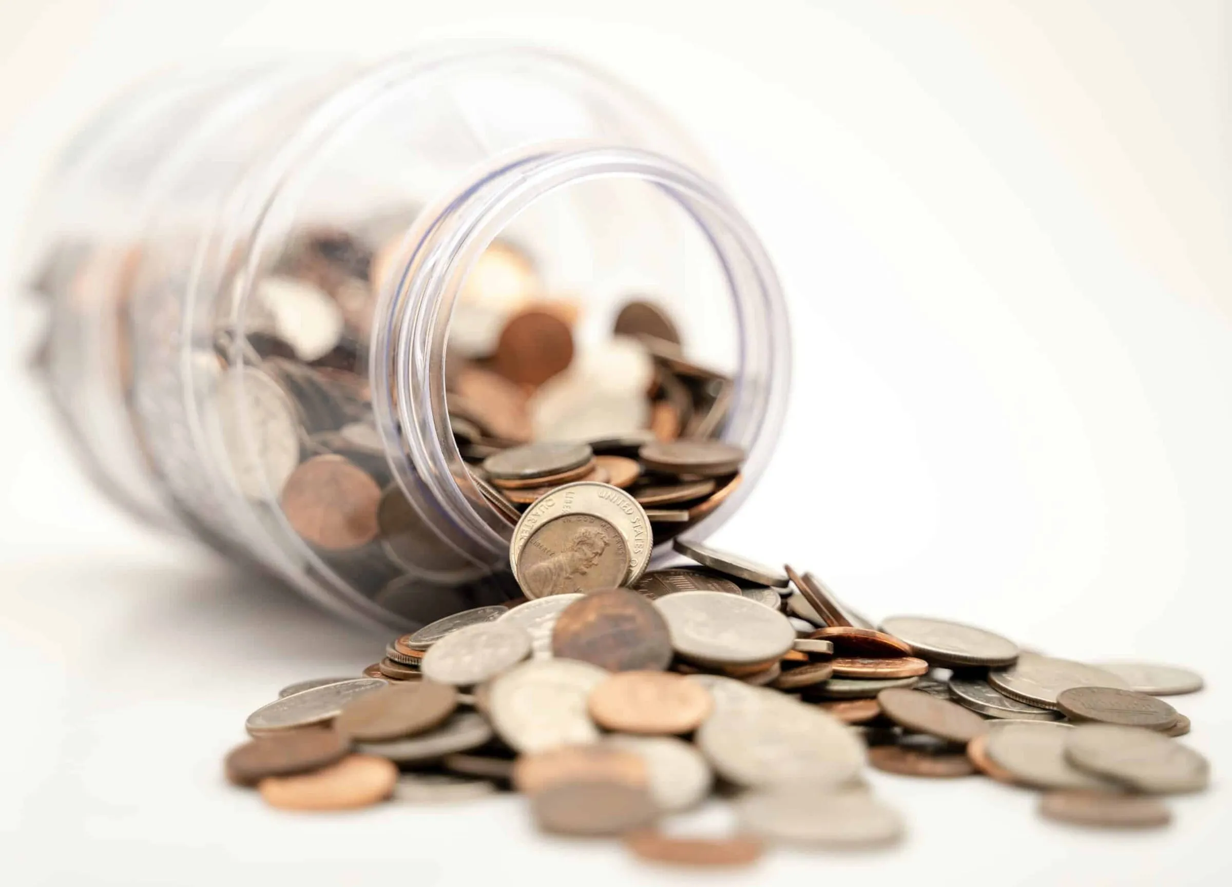 A glass jar tipped over with coins spilling out onto a white surface