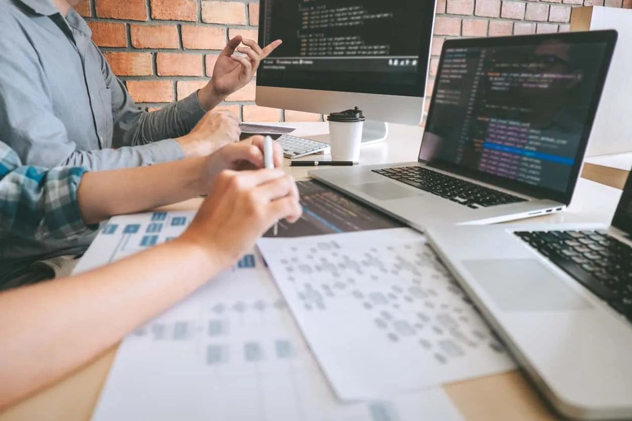 Two people sketching wireframes on paper beside a laptop showing code