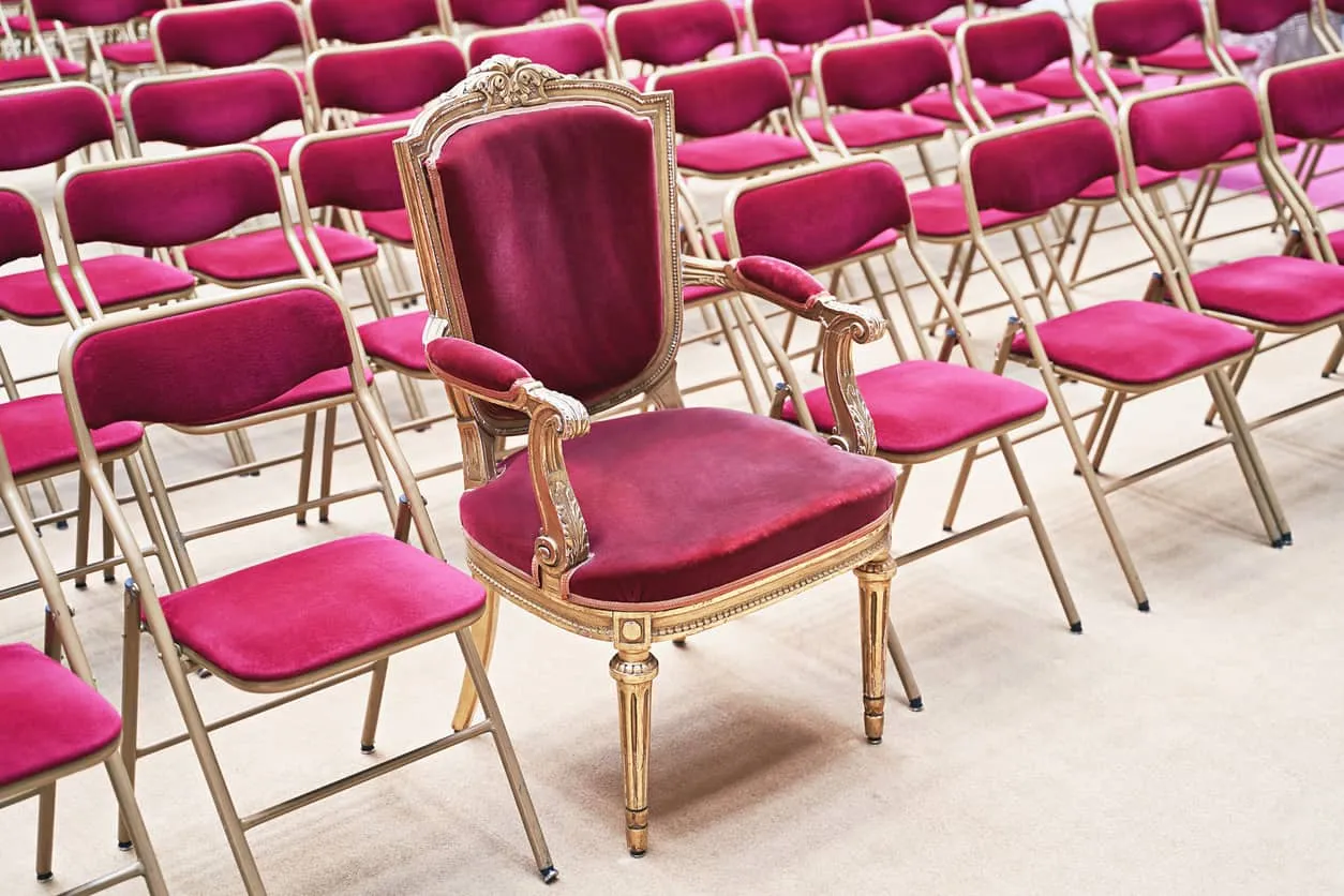 A single gilded armchair standing in front of rows of red banquet chairs