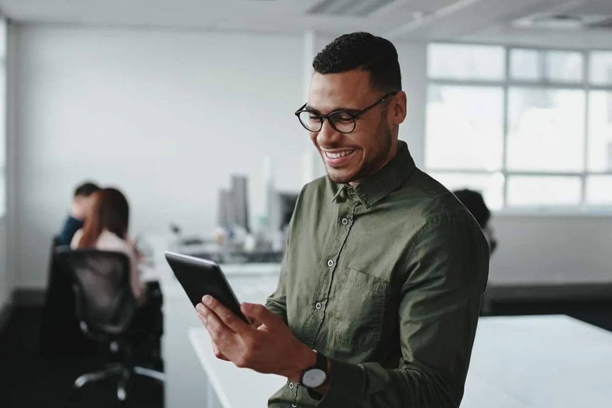 A man in glasses smiling at a tablet in an open plan office