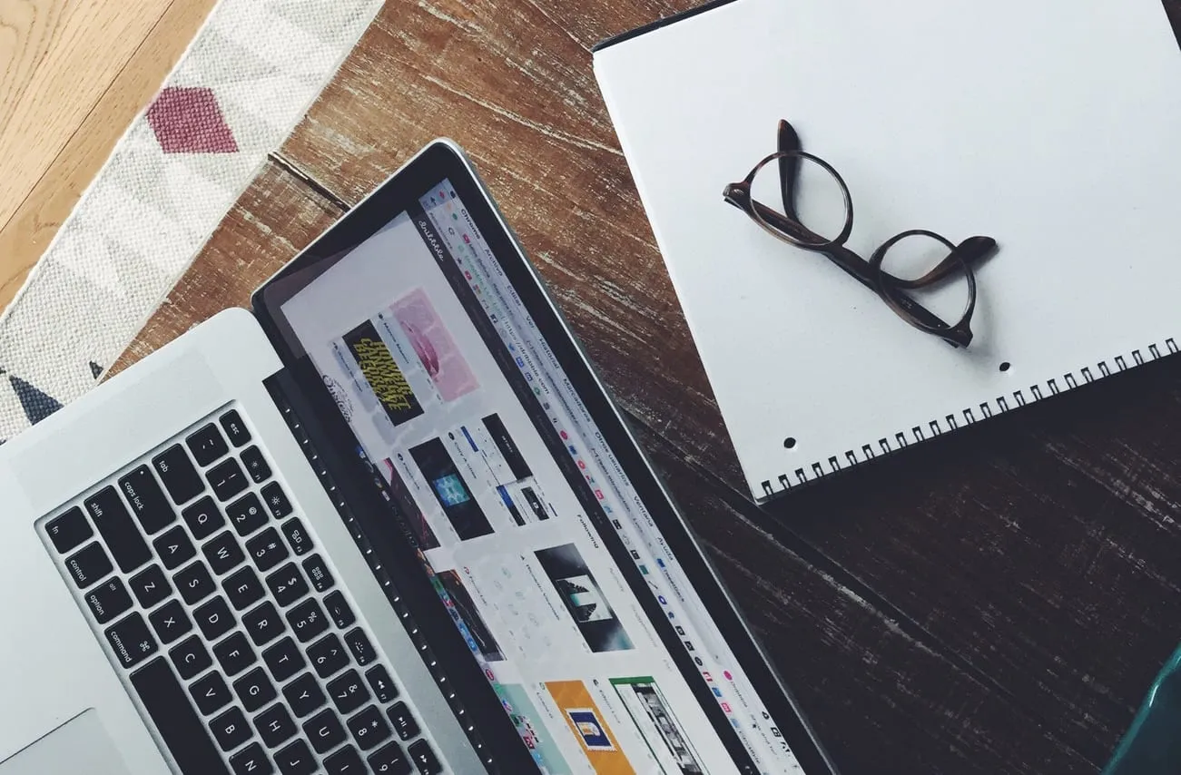 A laptop, notebook and pair of glasses on a wooden desk