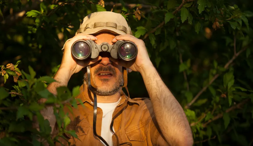 A man in a cap looking through binoculars from behind foliage