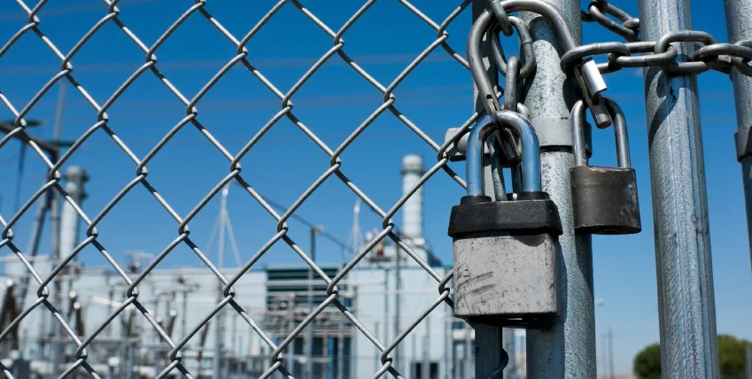 Two padlocks and a chain securing a wire fence gate in front of an industrial site