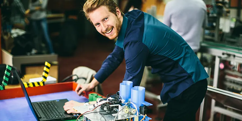 An engineer smiling at a laptop wired to a circuit board on a workbench