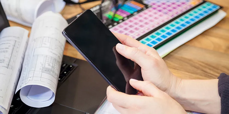 A hand holding a phone over a desk of rolled technical drawings and a keyboard
