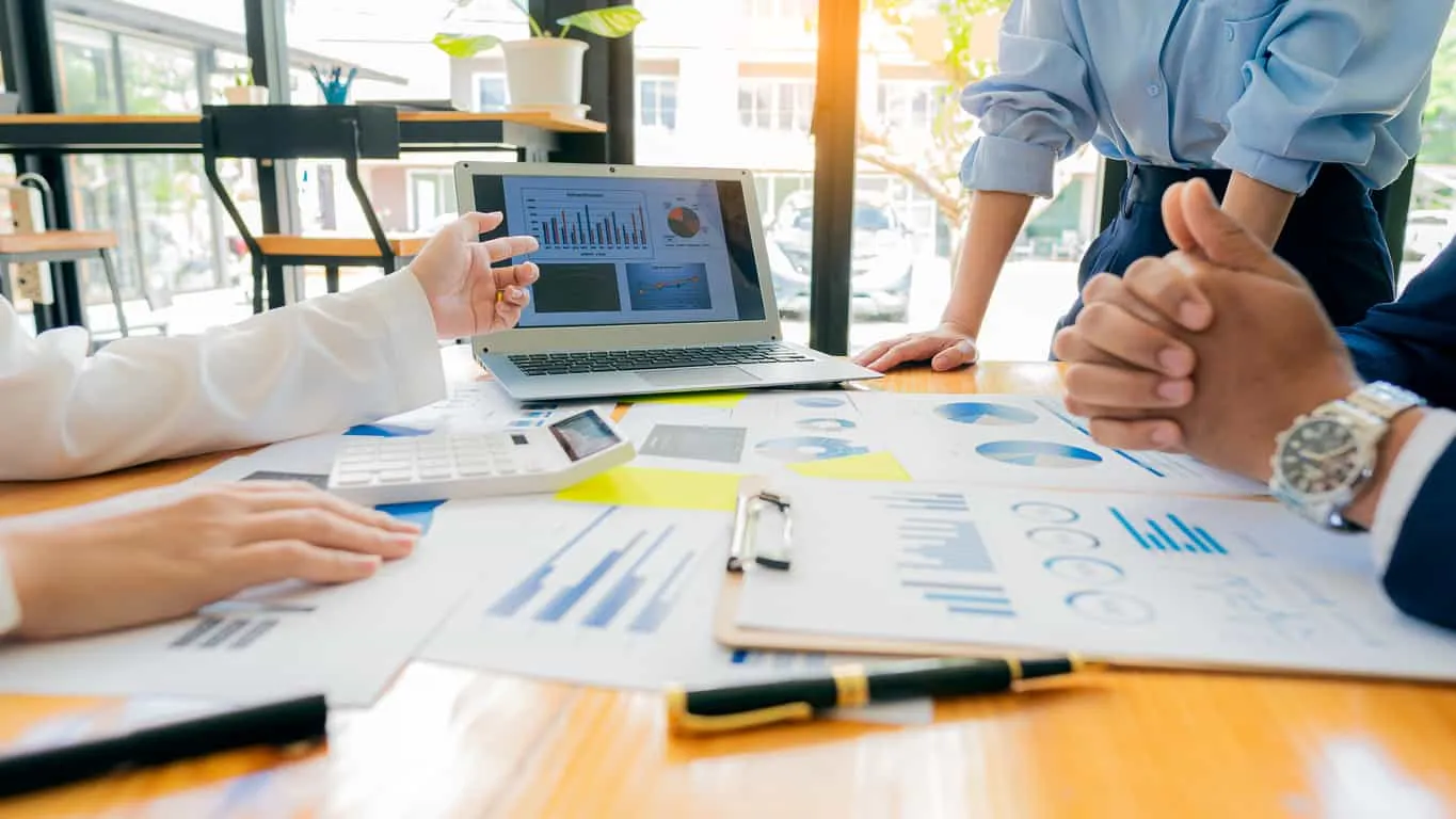 A team around a table of printed charts, one holding up a laptop