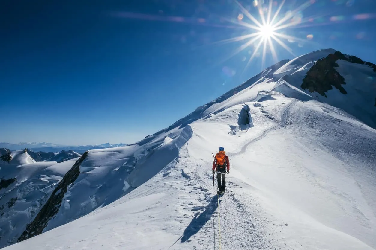 A climber walking a snow ridge towards a mountain summit in bright sun
