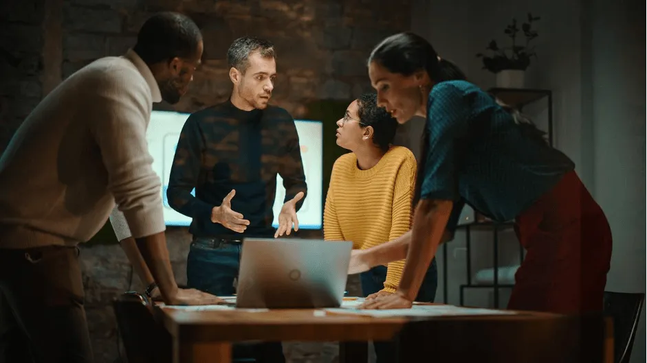 Four colleagues gathered around a laptop, one gesturing as he explains something