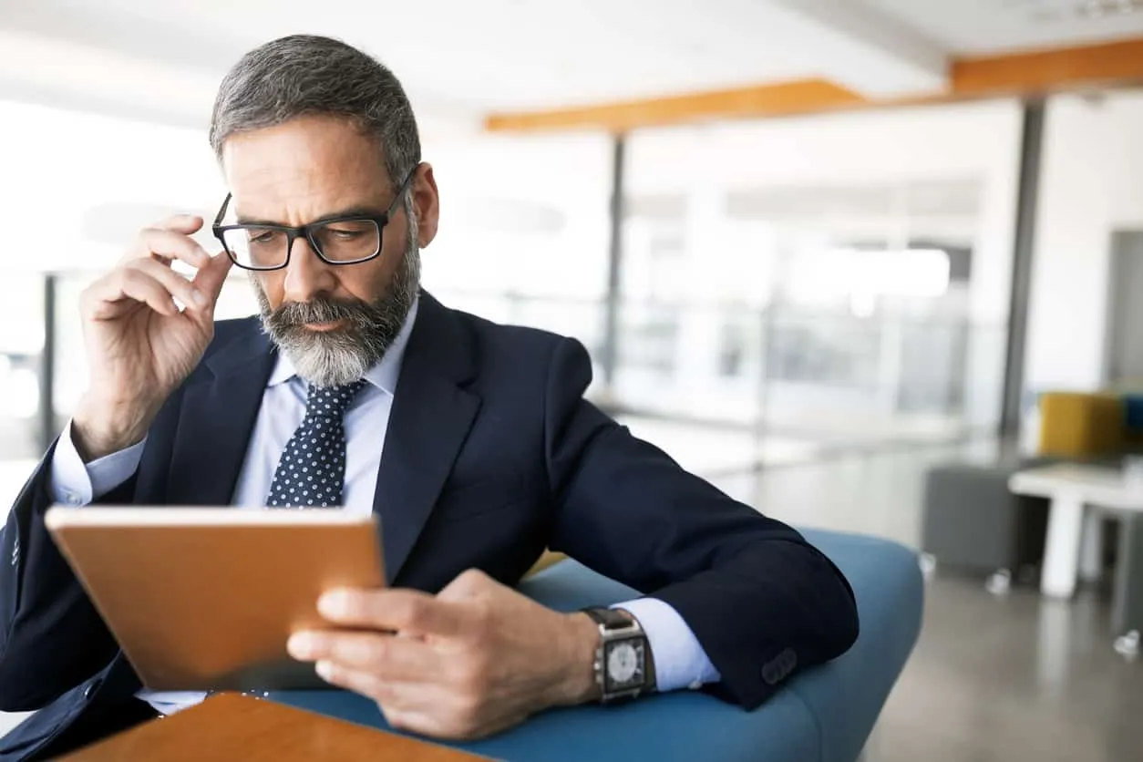 A man in a suit adjusting his glasses while reading a tablet