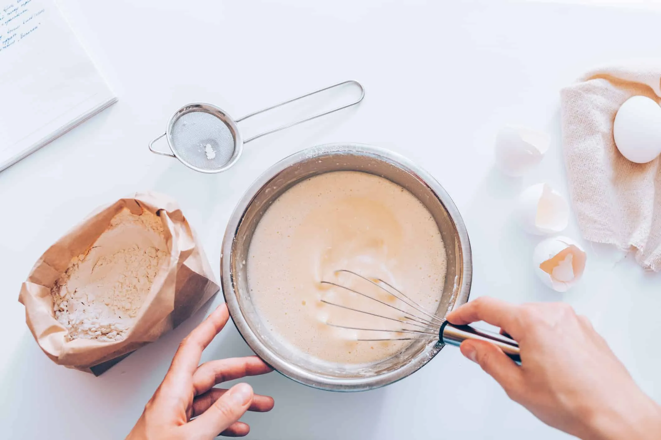 Overhead view of hands whisking batter in a bowl beside cracked eggshells and flour