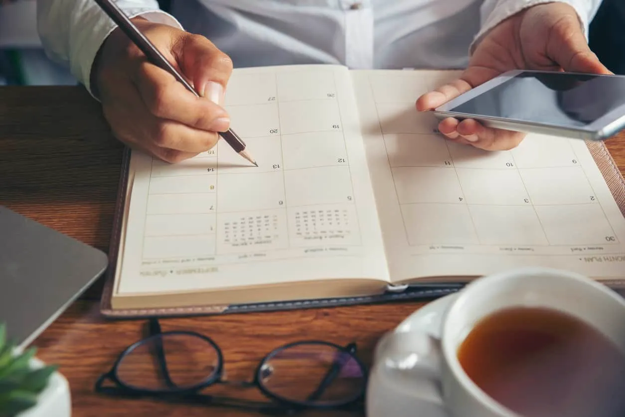 Hands writing in a paper planner beside a phone, glasses and a cup of tea