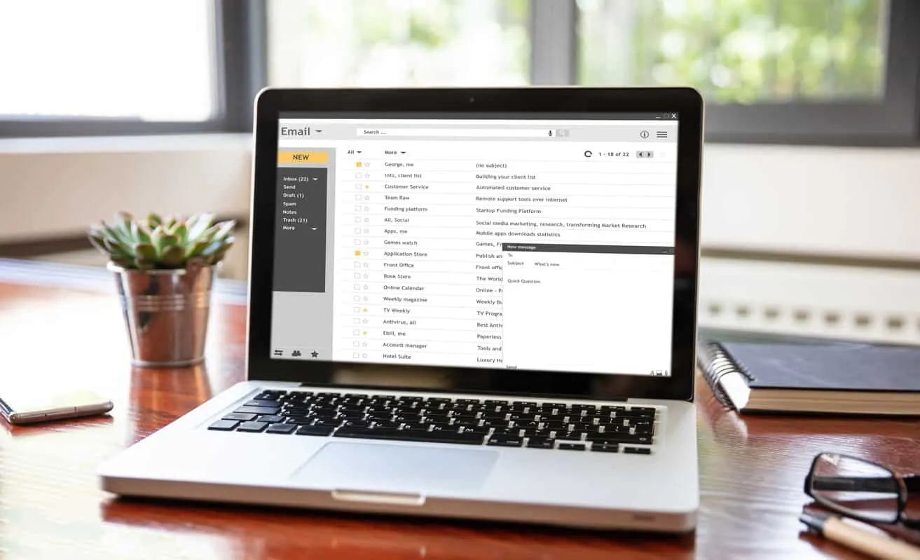 A laptop on a desk showing an email inbox, beside a small plant and a pair of glasses