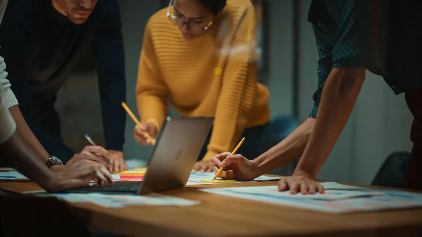 Three colleagues leaning over a table of notes and a laptop, working together