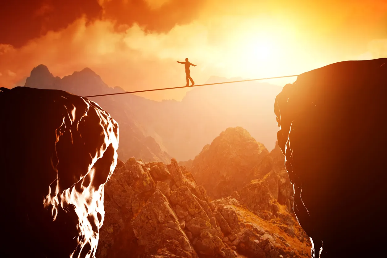 A person walking a tightrope strung between two mountain peaks at sunset