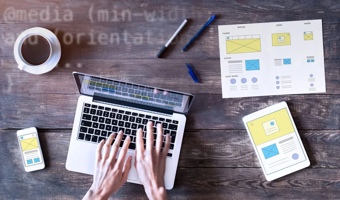 Overhead view of hands typing on a laptop surrounded by printed wireframes, a tablet and a phone