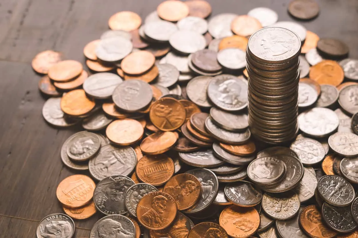 A stack of coins beside a scattered pile of loose change on a wooden surface