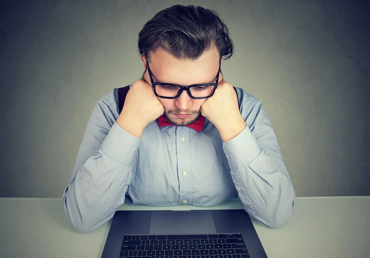 A man in glasses resting his chin on his hands, looking fed up in front of a laptop