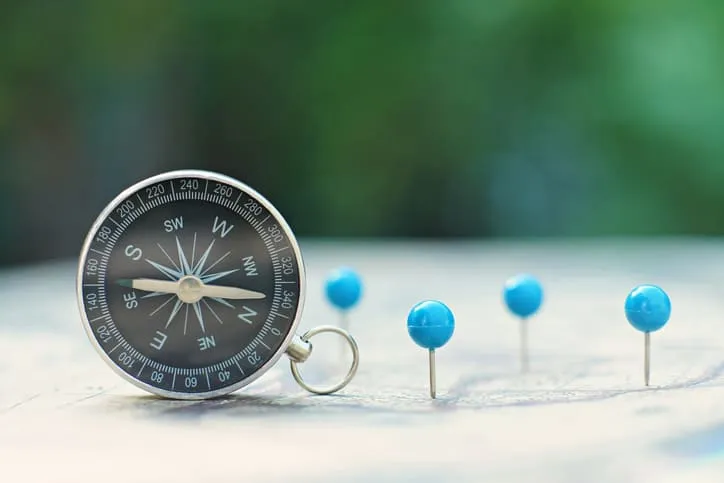 A compass on a sandy surface beside a row of blue map pins