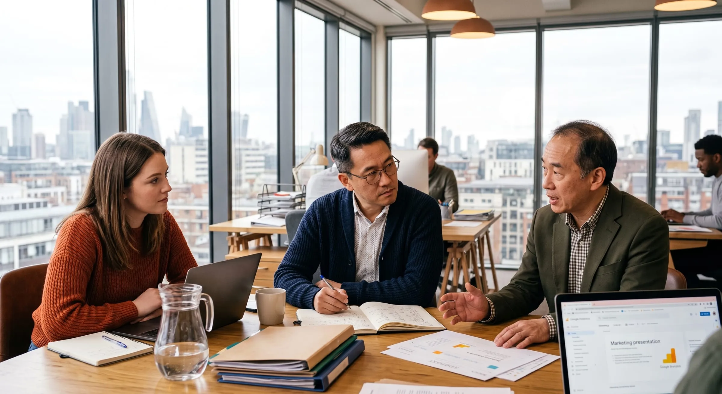 Three colleagues in discussion around a meeting table in a city-view office, notes and a laptop in front of them