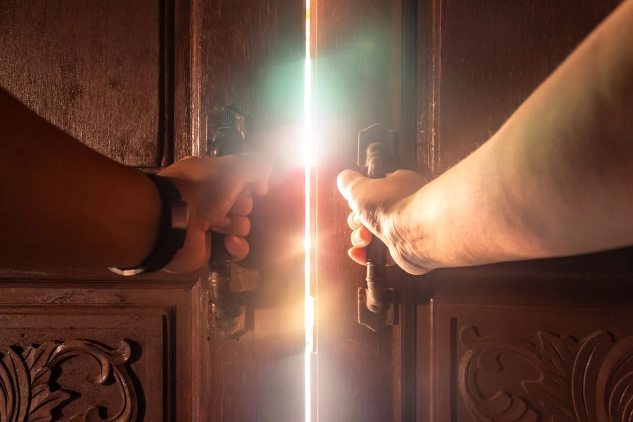 Hands pushing open a heavy wooden door with bright light behind it