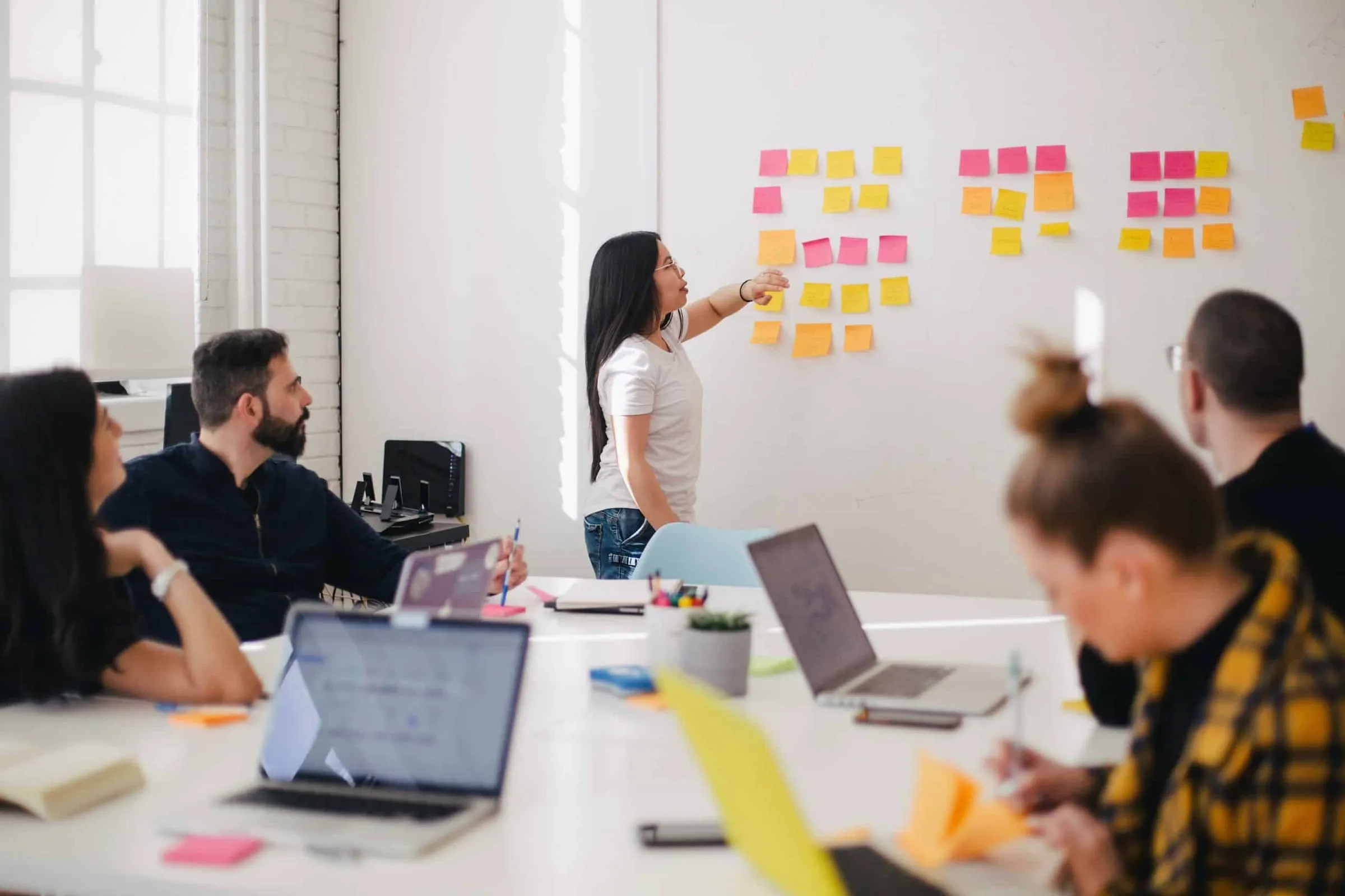A woman presenting at a wall of sticky notes to colleagues around a table