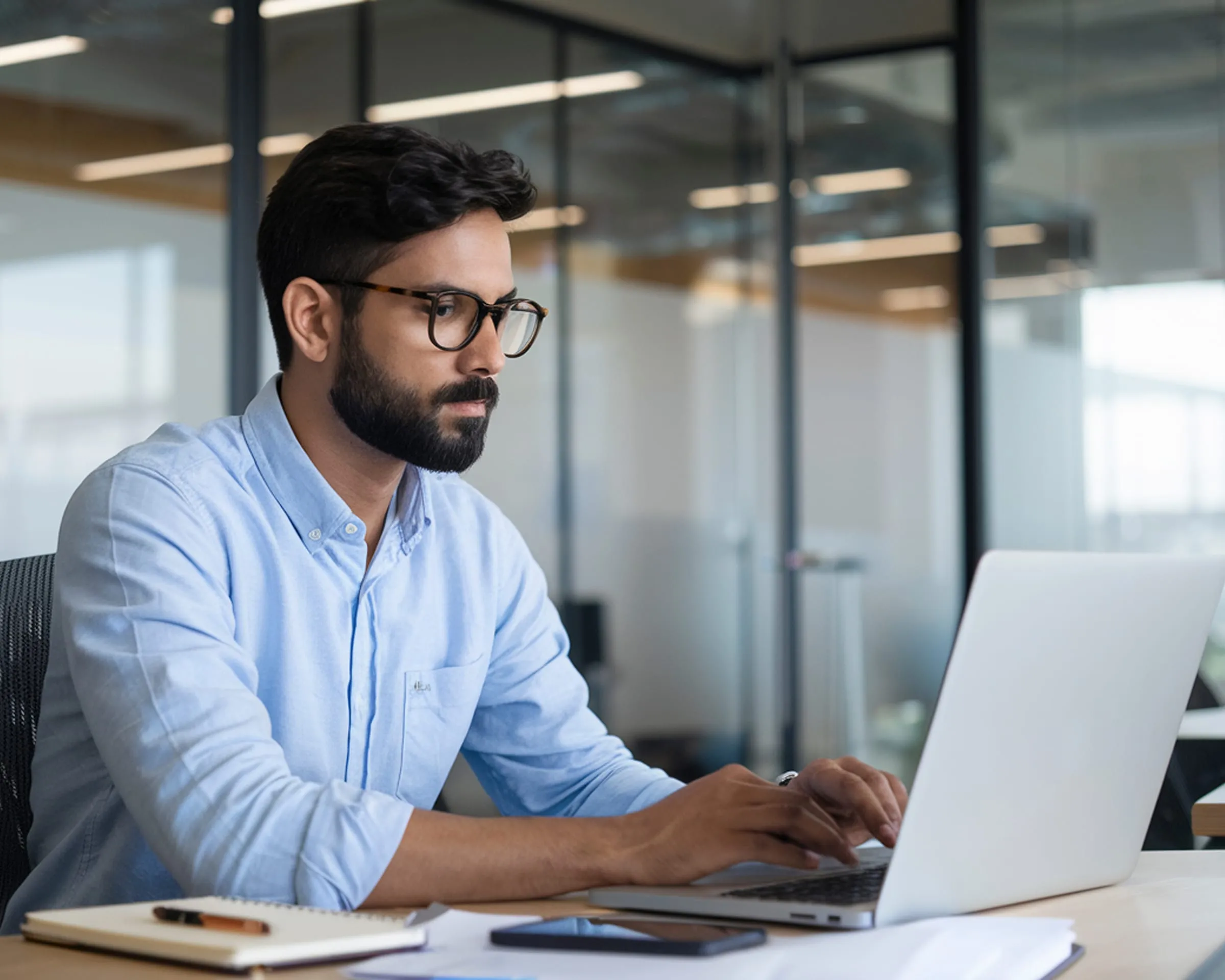 A man in glasses working at a laptop in a glass walled office