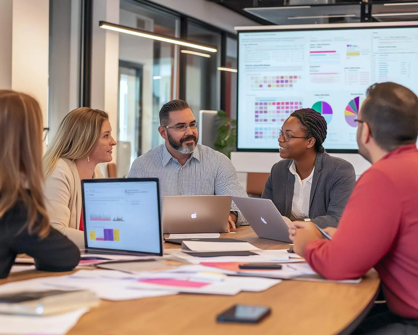 Five colleagues around a meeting table with laptops, watching a wall screen full of charts