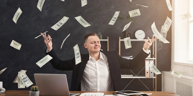 A man at a desk throwing his arms up as dollar bills fall around him