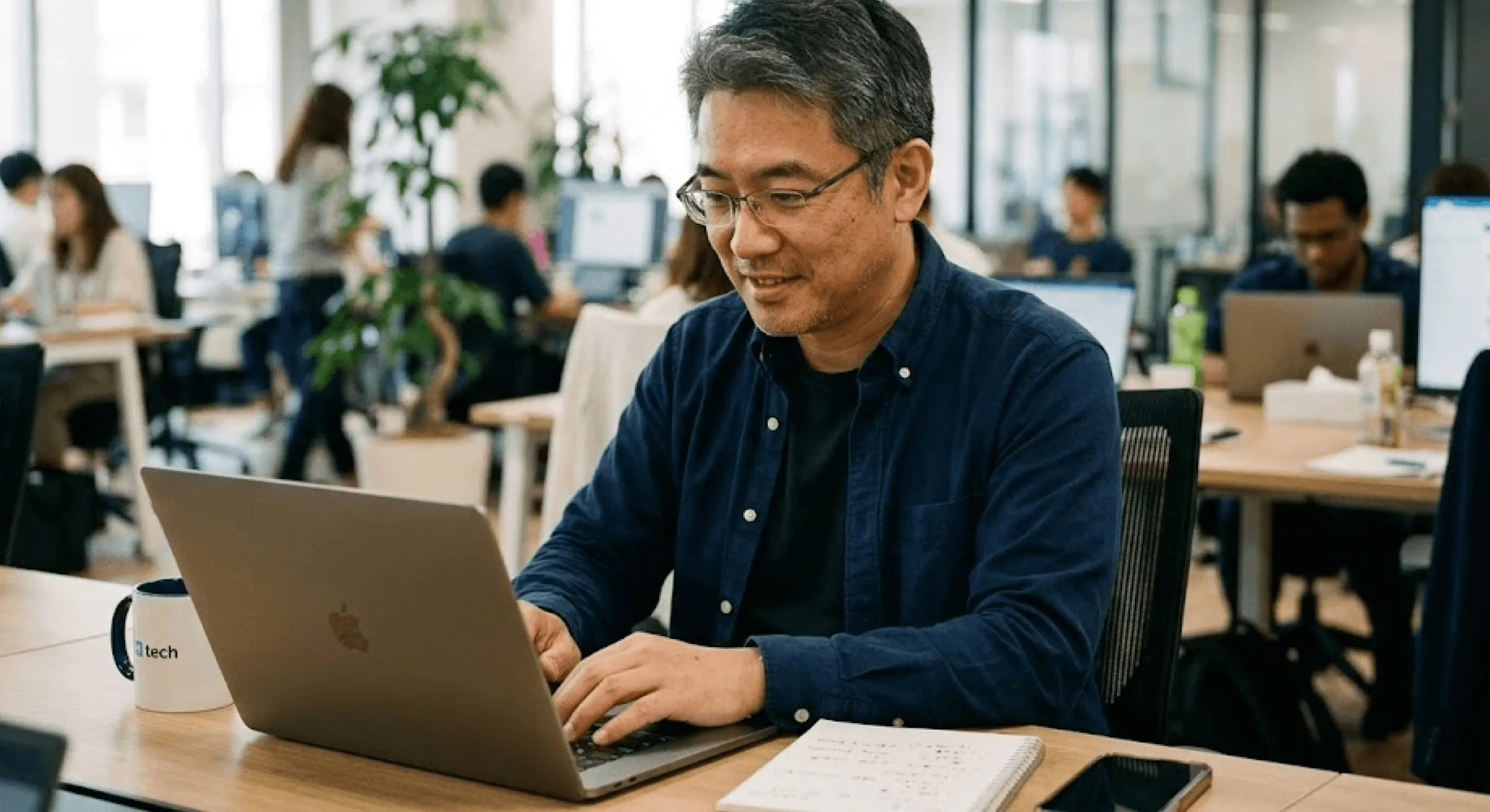 A person working at a laptop in a busy open-plan office, a notebook and coffee beside them
