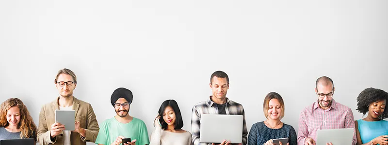 A row of people of different ages holding phones, tablets and laptops against a white wall