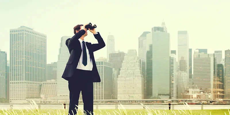 A man in a suit looking through binoculars with a city skyline behind him