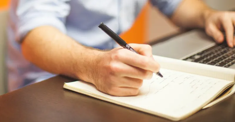 A man writing in a notebook beside an open laptop