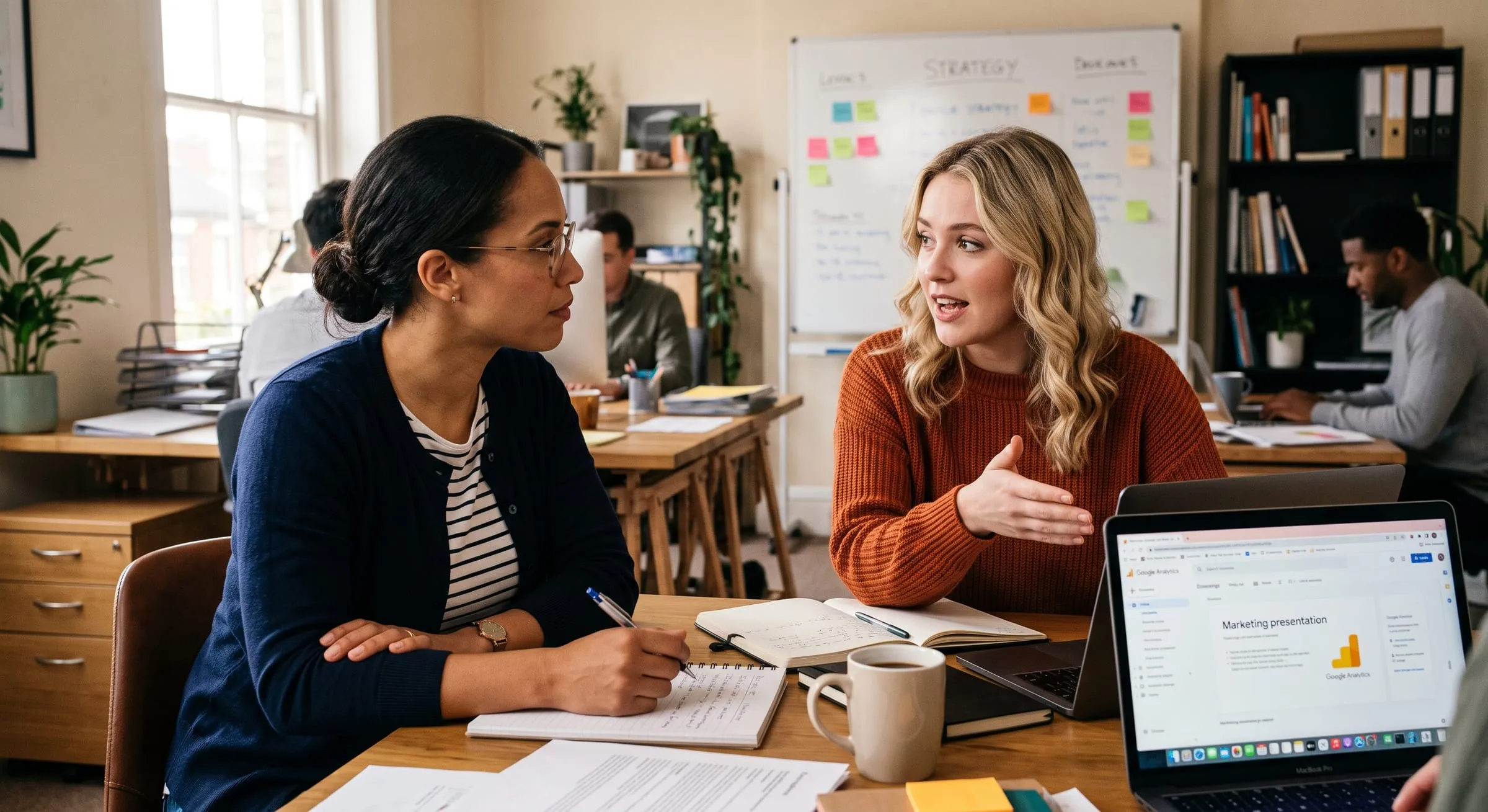 Two colleagues talking over notebooks and a laptop in an open-plan office, a strategy whiteboard behind them